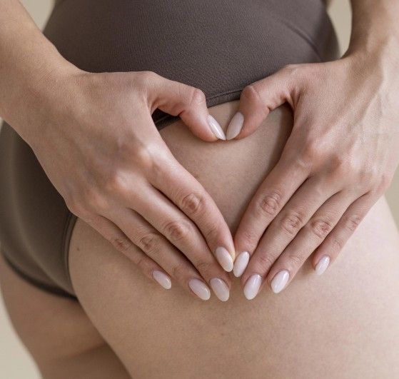 Primo piano del fianco di una donna in slip marrone: le mani, con unghie nude, formano un cuore poggiato sulla pelle.