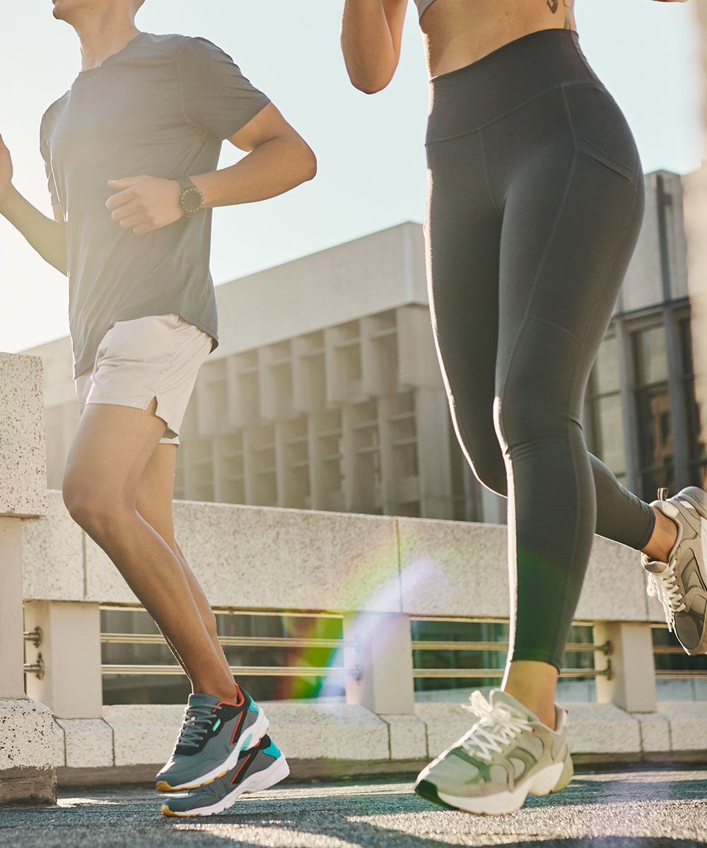 Due runner in abbigliamento sportivo che corrono su un marciapiede urbano, inquadrati su gambe e scarpe da ginnastica con luce solare.