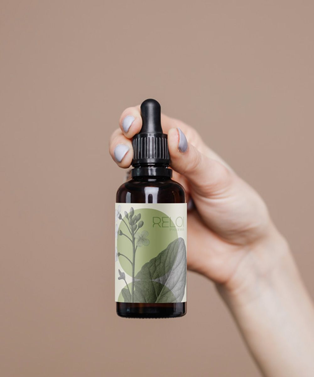 Primo piano di una mano che impugna un flacone ambrato di Kaïos Body Oil con etichetta bianca e verde decorata da illustrazioni botaniche.