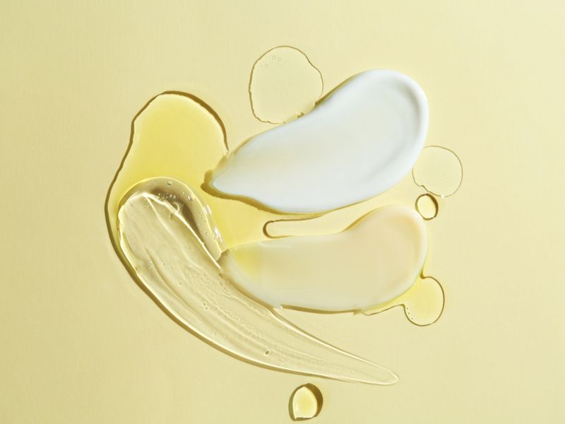 Texture cosmetiche su sfondo beige: crema bianca, olio dorato e gel trasparente disposti in composizione astratta.