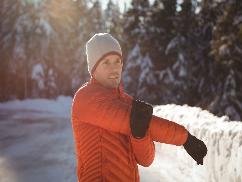 Persona con giacca arancione e berretto grigio che si riscalda allungando il braccio all’aperto in una giornata invernale, con neve e alberi sullo sfondo.