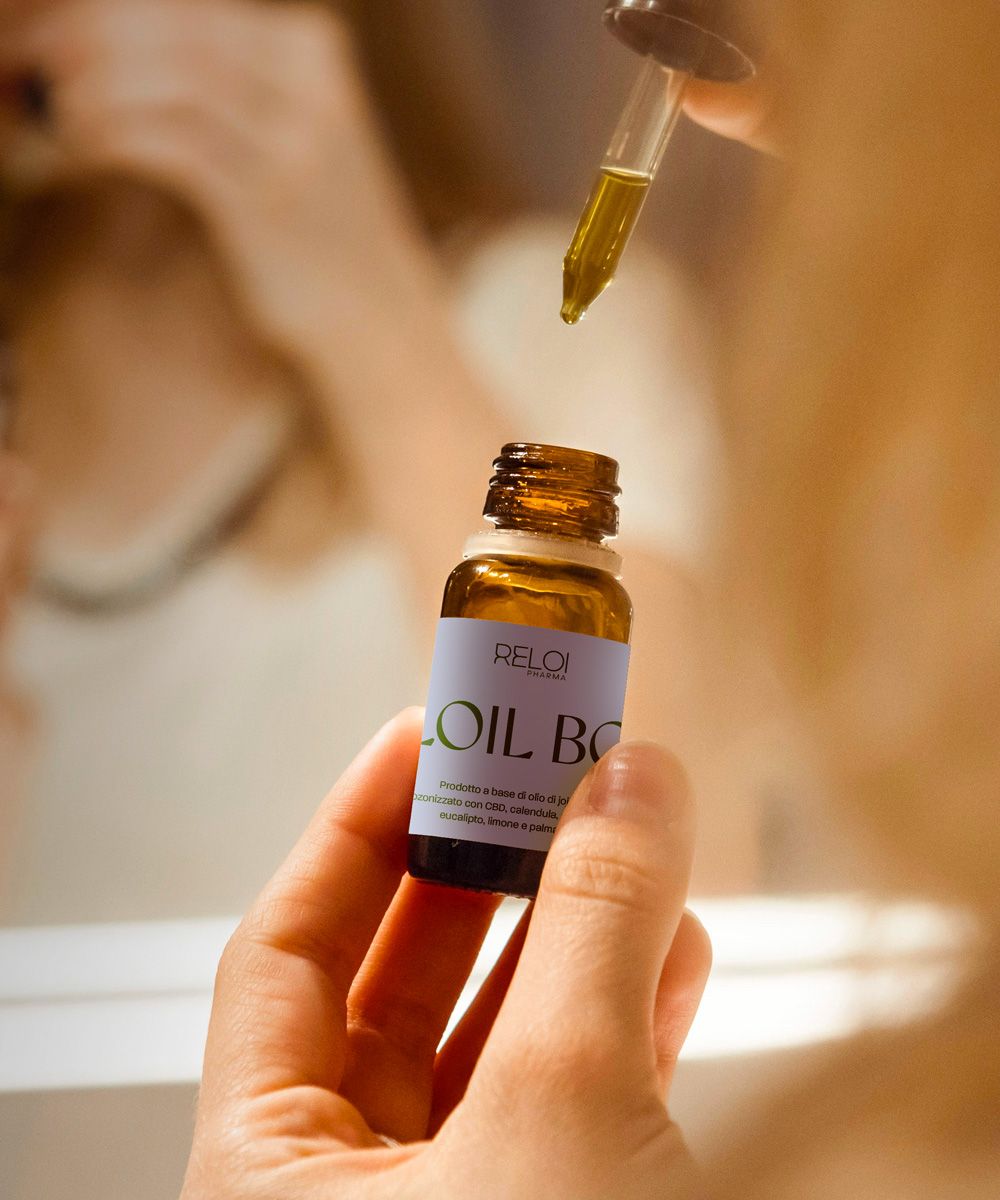 Primo piano di mani che versano poche gocce di Reloil Body Oil da un contagocce su un flacone in vetro ambrato, sfondo bagno sfocato.