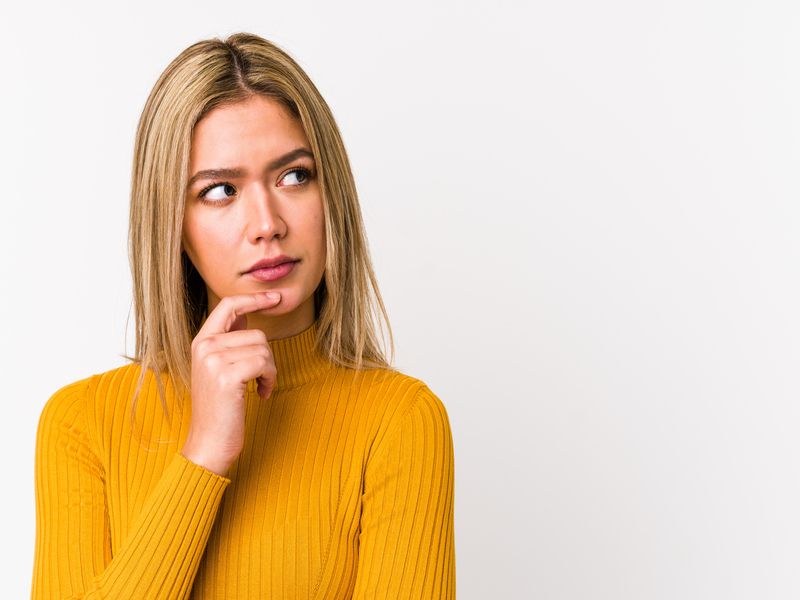 Donna con maglione senape che guarda di lato con espressione pensierosa, su sfondo neutro chiaro