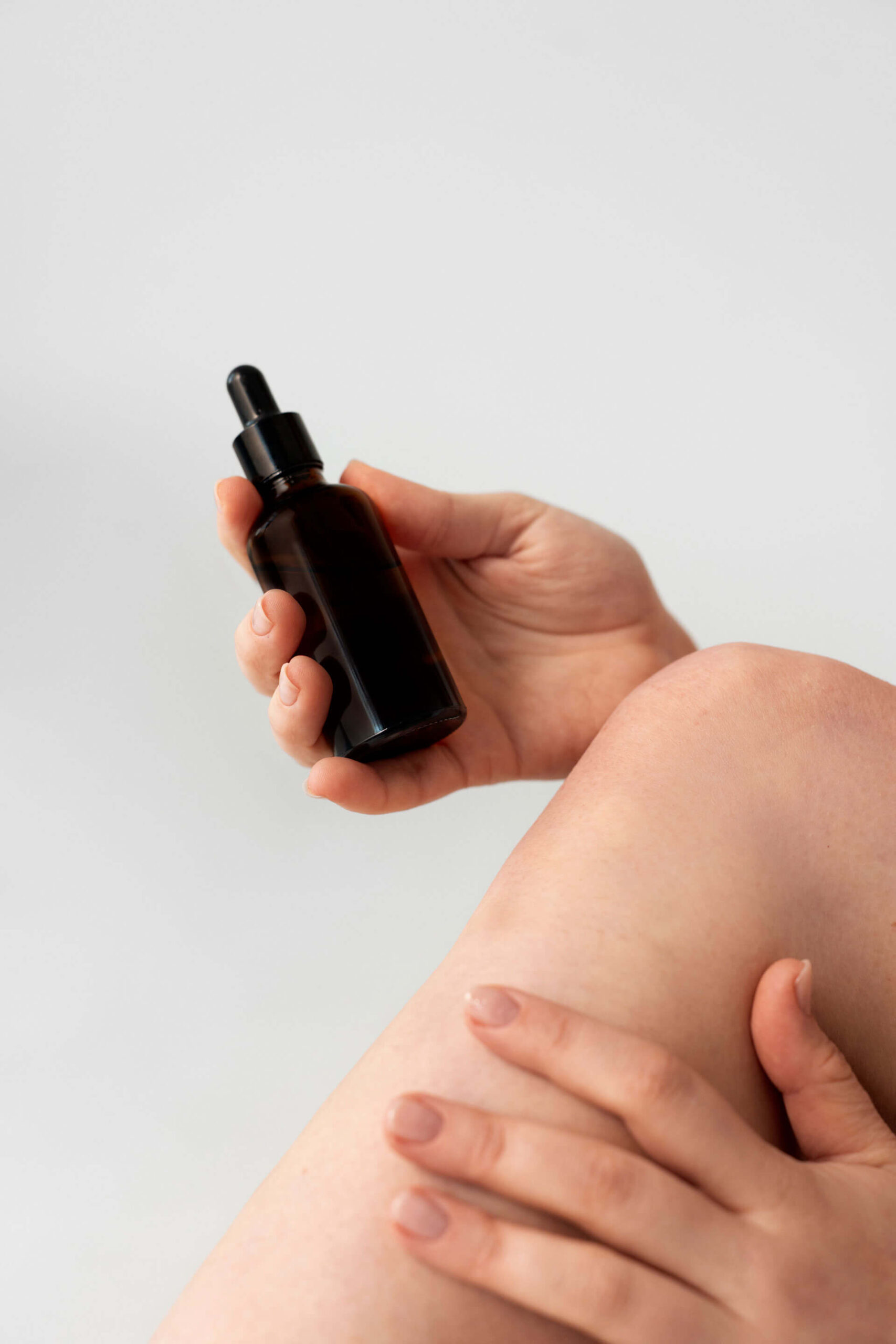 Mano che tiene un flacone di vetro scuro con contagocce mentre lo applica su una gamba, su sfondo neutro.