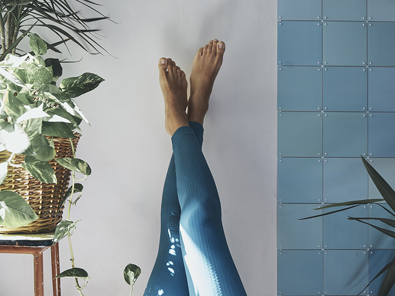 Donna in leggings solleva le gambe contro il muro per favorire il microcircolo e il defaticamento muscolare,, in un ambiente rilassante con luce naturale e piante