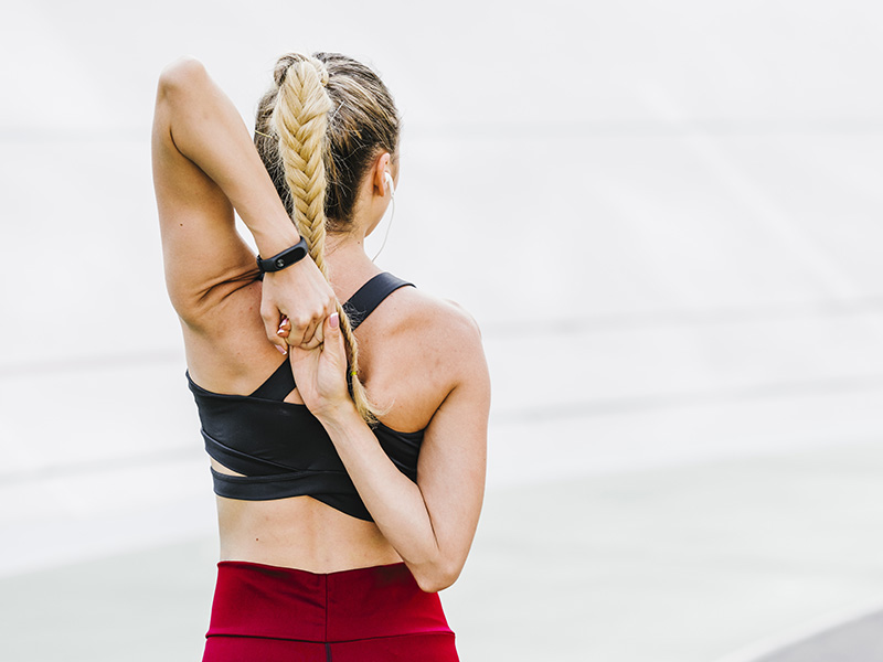 Donna sportiva fa stretching delle spalle all’aperto, vista da dietro.