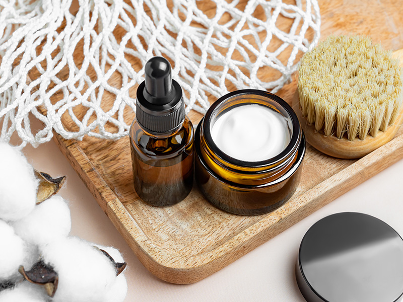 Crema rassodante e olio corpo in vasetto e boccetta su vassoio in legno, con accessori skincare