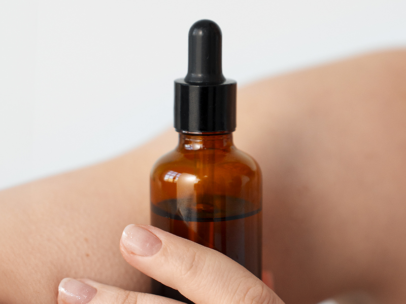 Mano che tiene boccetta in vetro scuro Miron con contagocce, olio corpo Reloil.