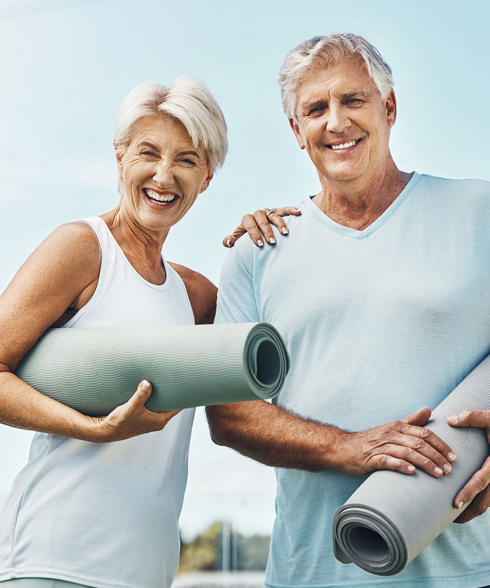 Coppia di senior sorridenti in abbigliamento sportivo che tengono tappetini da yoga, sfondo cielo sereno