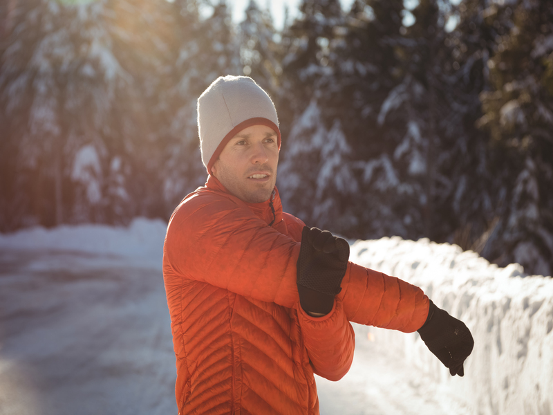 Persona con giacca arancione e berretto grigio che si riscalda allungando il braccio all’aperto in una giornata invernale, con neve e alberi sullo sfondo.