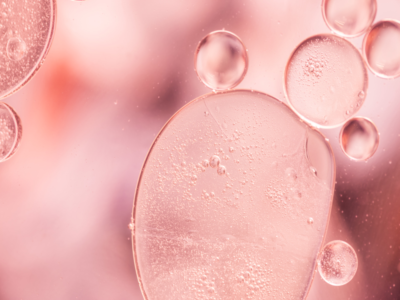 Macrofotografia di bolle d’olio rosa sospese in liquido, con forme rotonde e traslucide che si sovrappongono in un effetto morbido.