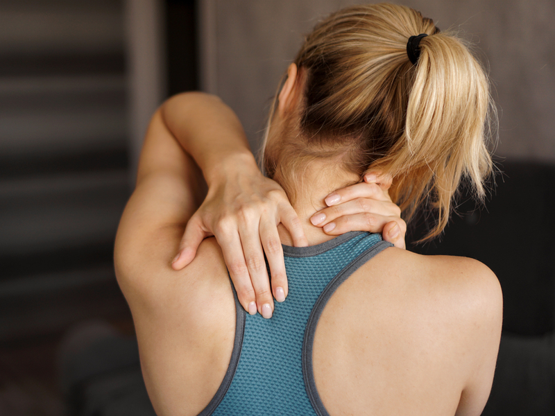 Donna sportiva di spalle si massaggia il collo e la spalla dopo un allenamento, con espressione di dolore o tensione muscolare.