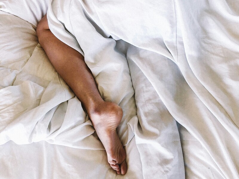 Piedi nudi di una persona distesa a letto, parzialmente coperta da lenzuola bianche.