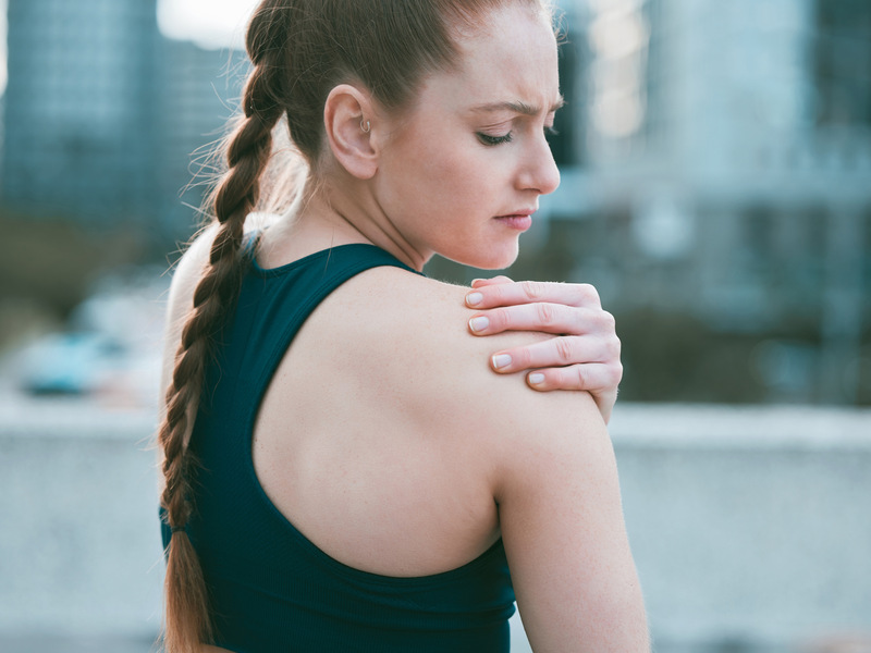 Donna in abbigliamento sportivo che si tiene la spalla dolorante durante un allenamento all’aperto, con espressione di fastidio.