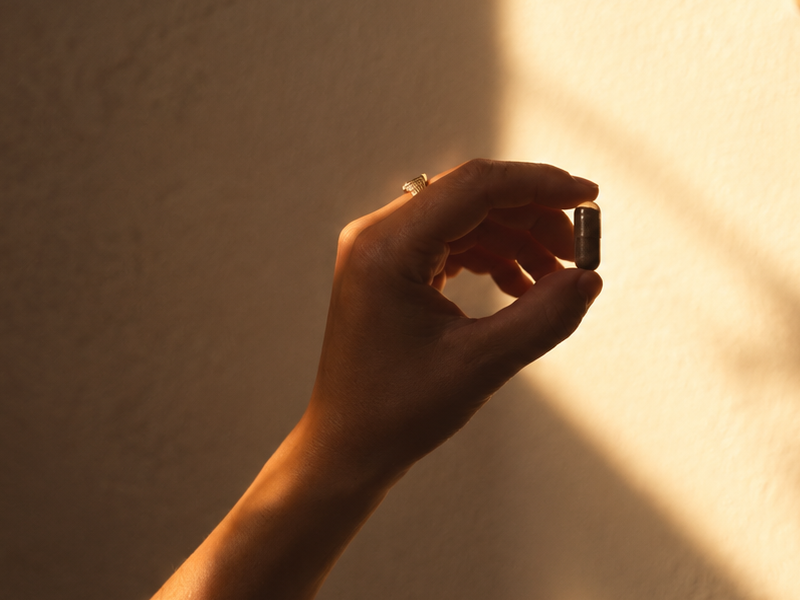 Mano illuminata dalla luce calda del sole che tiene una piccola capsula, in un ambiente interno dai toni neutri e con ombre morbide.